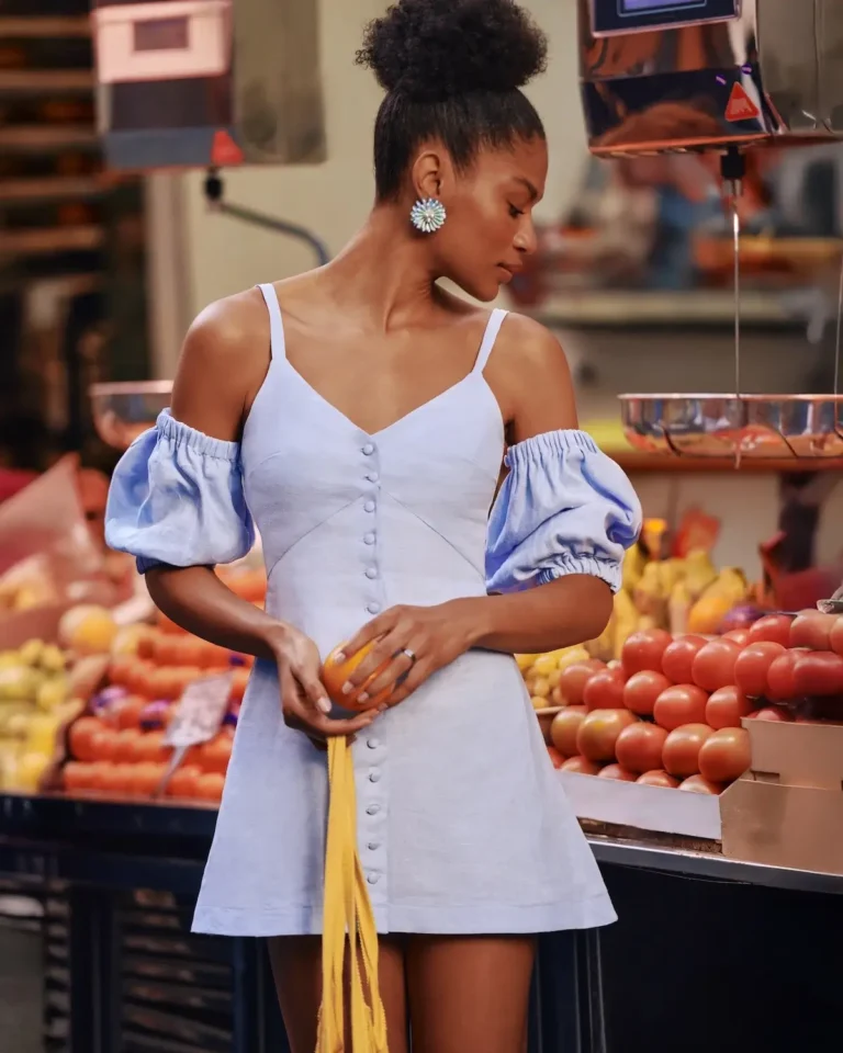 Model wearing blue linen button down mini dress with detachable puff sleeves in market setting.